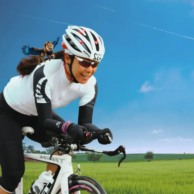 Radfahrerin im Zeitfahrhelm auf einem Rennrad vor grüner Wiese, auf ihrer Schulter ein geflügelter Löwe