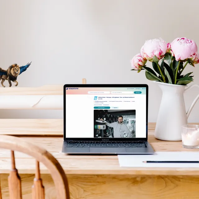 Laptop auf Holztisch zeigt eine Stellenanzeige, daneben ein Blumenstrauß und ein kleiner geflügelter Löwe